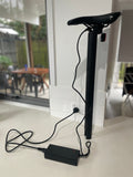 The seat post stem battery being charged on a kitchen bench. The charger is plugged into the battery and wall and there is a table in chairs blurred out in the background.